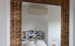 Woven rattan mirror frame reflecting a cozy bedroom with a colorful fish-patterned headboard and decorative pillows, featuring a wall-mounted air conditioner in a bright, minimalist space.