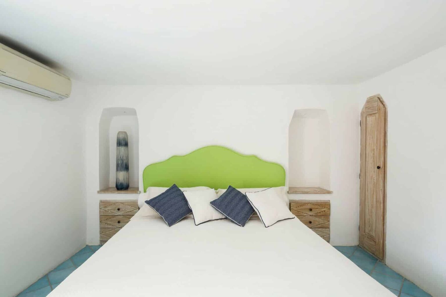 Modern bedroom featuring a white color scheme, a green headboard, and blue accents. The room includes two wooden nightstands with decorative pillows on the bed and a wall-mounted air conditioning unit. | Only on SopranoVillas