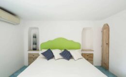 Modern bedroom featuring a white color scheme, a green headboard, and blue accents. The room includes two wooden nightstands with decorative pillows on the bed and a wall-mounted air conditioning unit.