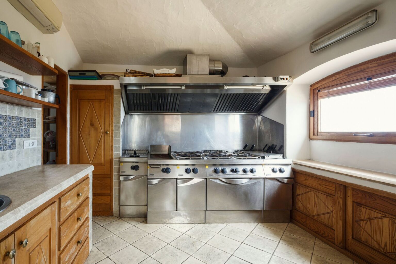 Modern kitchen interior featuring stainless steel appliances, a professional-grade cooking range, and wooden cabinetry. The space is well-lit with natural light from a window, showcasing a clean and functional design. | Only on SopranoVillas