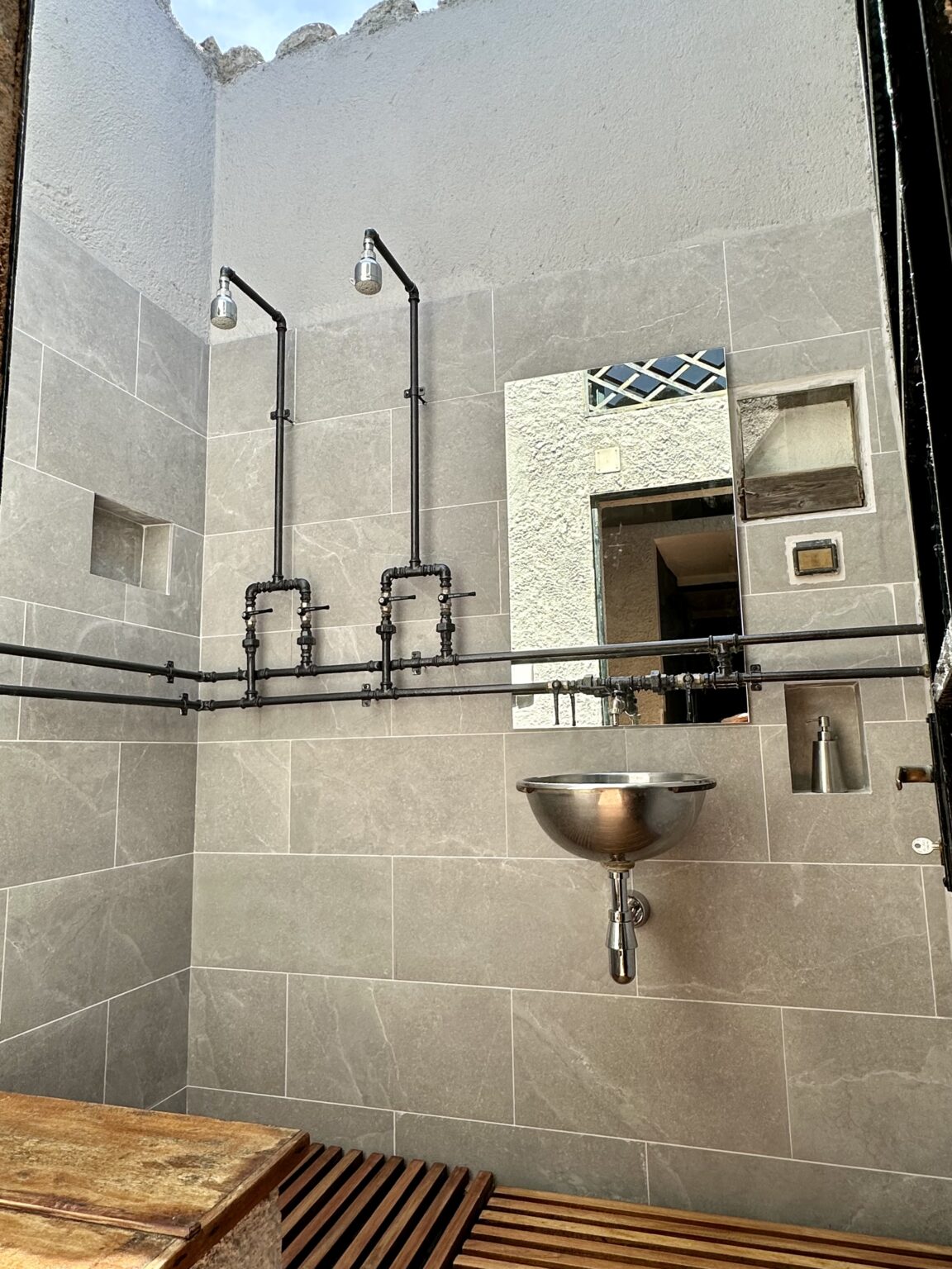 Modern industrial bathroom design featuring gray tiled walls, dual showerheads, a round metal sink, and wooden slatted flooring. The space is illuminated by natural light from an open ceiling, highlighting the minimalist aesthetic. | Only on SopranoVillas