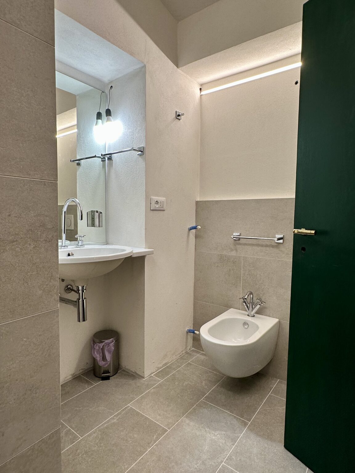 Modern bathroom featuring a wall-mounted sink, bidet, and sleek gray tile flooring. The space is illuminated by pendant lights and includes a green door, enhancing the contemporary design. | Only on SopranoVillas