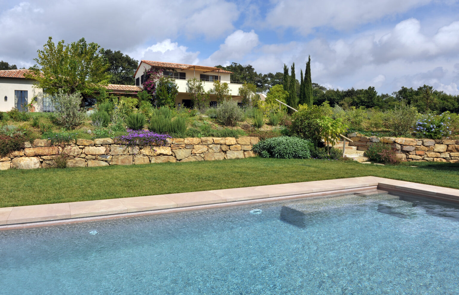 A serene backyard view featuring a clear swimming pool, lush greenery, and a beautifully landscaped garden with stone walls and colorful flowers, set against a backdrop of a modern home and blue sky. | Only on SopranoVillas