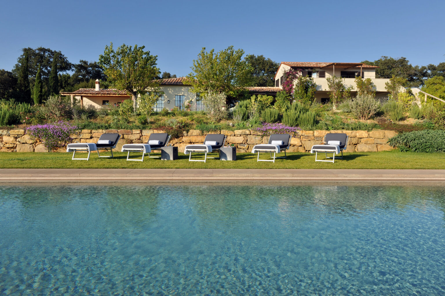 Luxurious outdoor pool area featuring modern lounge chairs, lush greenery, and a charming villa in the background under a clear blue sky. Perfect for relaxation and summer gatherings. | Only on SopranoVillas
