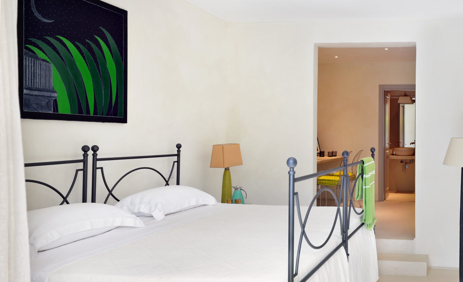 Cozy bedroom featuring a black metal bed frame with white linens, a bedside lamp, and a vibrant green art piece on the wall. The room has a warm, minimalist design with an open doorway leading to a modern bathroom. | Only on SopranoVillas