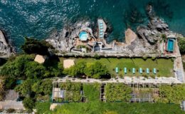 Aerial view of a coastal property featuring swimming pools, rocky shoreline, and lush greenery. The image showcases the vibrant blue water, landscaped gardens, and lounging areas, highlighting a luxurious outdoor retreat by the sea.