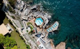 Aerial view of a luxurious seaside villa with a private pool, surrounded by rocky cliffs and clear blue water, featuring sun loungers and landscaped gardens.