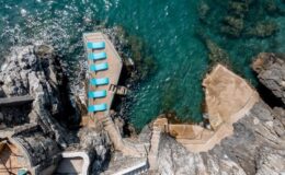 Aerial view of a rocky coastline featuring a pathway leading to a small dock with blue lounge chairs overlooking clear turquoise waters.