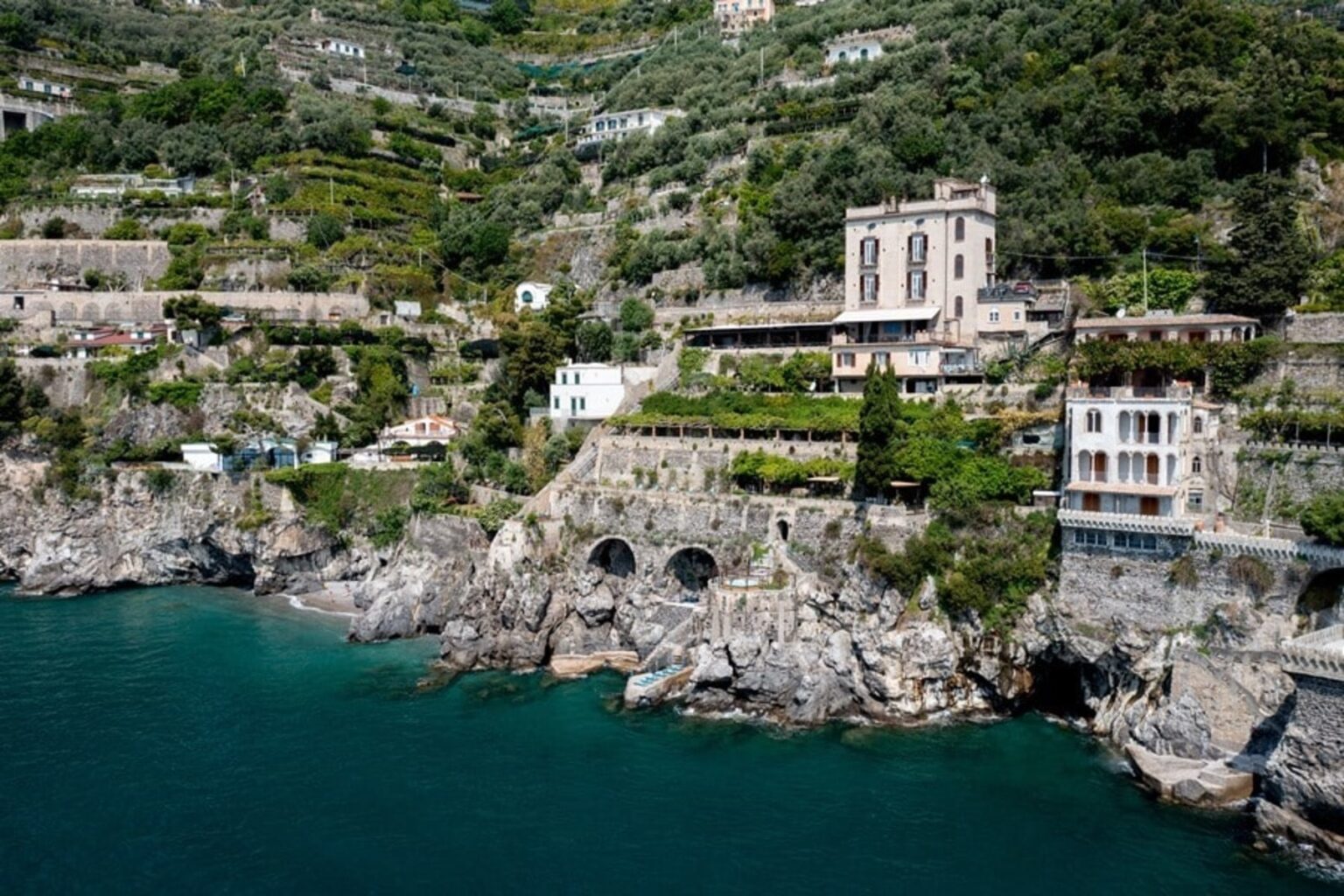 Coastal view of luxury villas and terraced gardens along the cliffs of the Amalfi Coast, Italy, with clear turquoise waters below. | Only on SopranoVillas