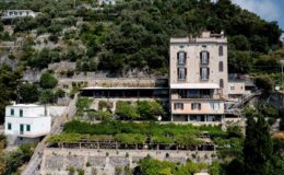 Aerial view of a hillside villa with terraced gardens and surrounding greenery, showcasing Mediterranean architecture and scenic landscape.