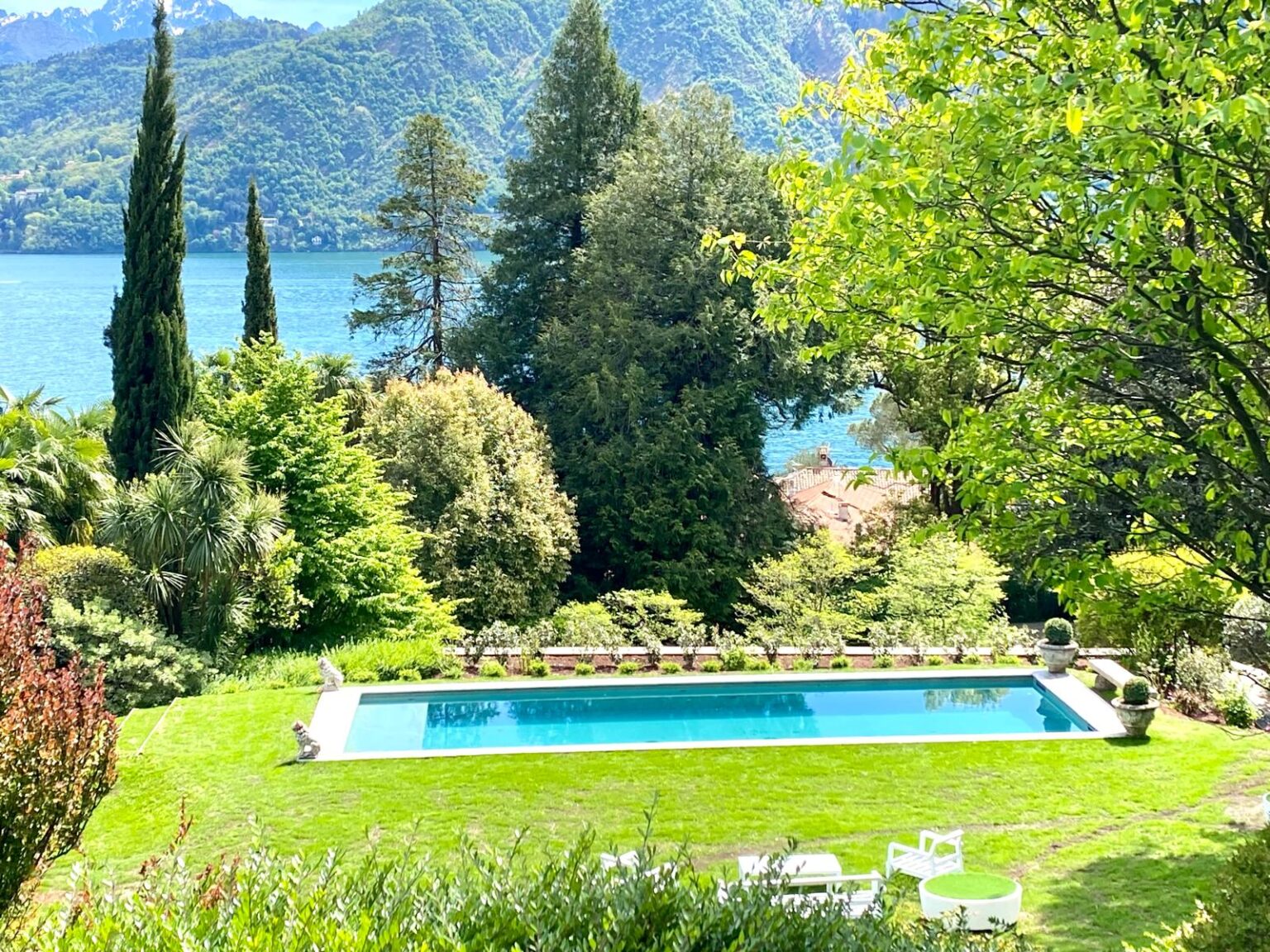 Scenic view of a lush garden with a swimming pool overlooking a serene lake, surrounded by mountains and greenery, ideal for relaxation and outdoor activities. | Only on SopranoVillas