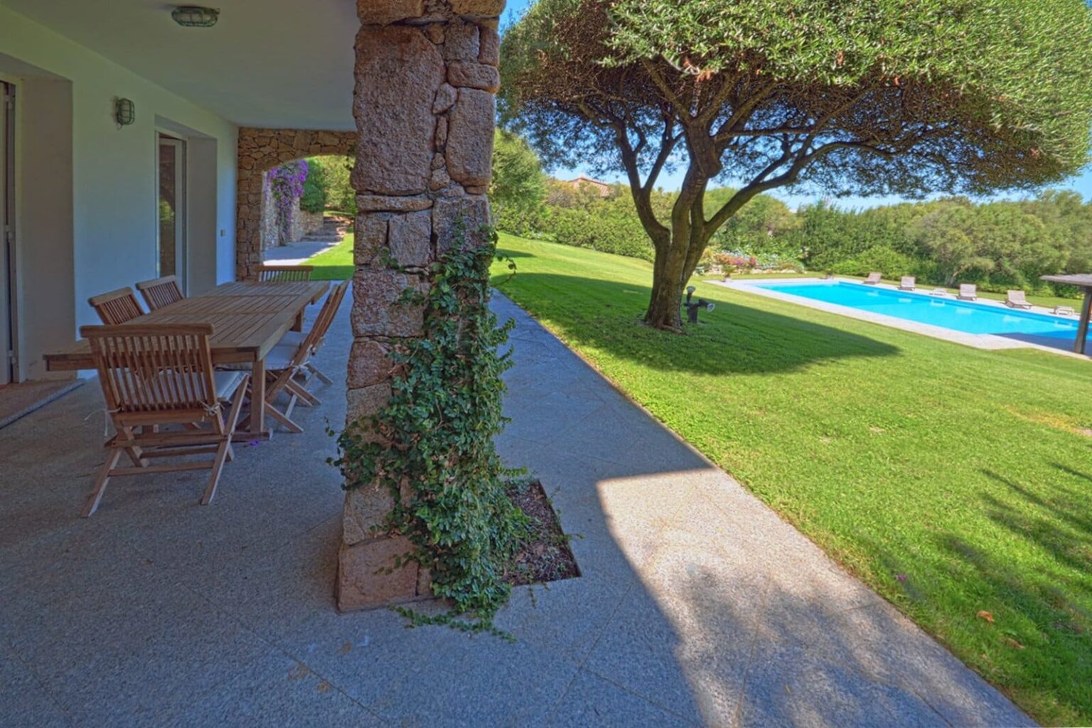 Modern outdoor patio with a wooden dining table, overlooking a lush green lawn and a swimming pool surrounded by trees. Ideal for relaxation and outdoor dining. | Only on SopranoVillas