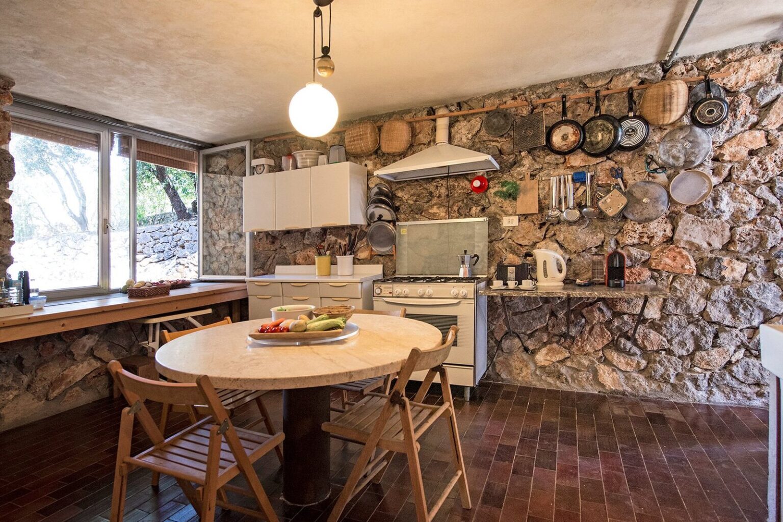 Cozy kitchen featuring a rustic stone wall, wooden dining table with fresh fruit, and modern appliances, illuminated by a hanging light fixture. | Only on SopranoVillas