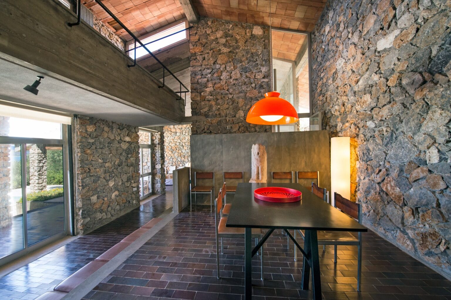 Modern dining area featuring stone walls, a sleek black table, wooden chairs, and a bright orange pendant lamp, showcasing contemporary architecture and design aesthetics. | Only on SopranoVillas