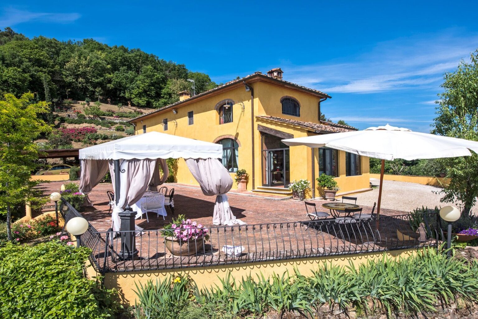 Beautiful yellow villa with outdoor seating area and white umbrellas, surrounded by lush greenery and blue sky, ideal for relaxation and scenic views. | Only on SopranoVillas
