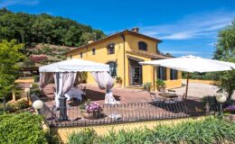 Beautiful yellow villa with outdoor seating area and white umbrellas, surrounded by lush greenery and blue sky, ideal for relaxation and scenic views.