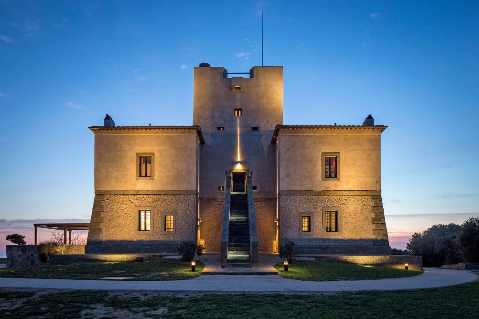 Historic stone castle illuminated at dusk, featuring a modern entrance and surrounded by landscaped grounds, showcasing architectural grandeur and charm. | Only on SopranoVillas