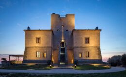 Historic stone castle illuminated at dusk, featuring a modern entrance and surrounded by landscaped grounds, showcasing architectural grandeur and charm.