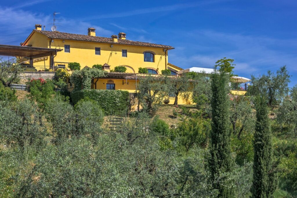 Vibrant yellow villa surrounded by lush olive trees under a clear blue sky, showcasing a picturesque countryside setting. | Only on SopranoVillas