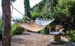 Hammock tied between two trees in a serene outdoor setting, with a rustic wooden table holding a glass of wine and a laptop, surrounded by lush greenery and a view of the ocean.