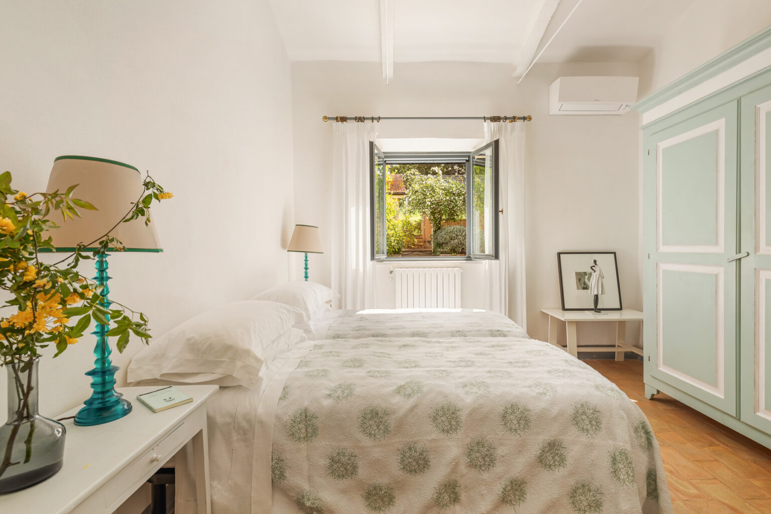 Bright and airy bedroom featuring a cozy double bed with a patterned duvet, a bedside table with a teal lamp, and a window overlooking a garden. The room has light-colored walls, wooden flooring, and a vintage-style wardrobe, creating a welcoming and serene atmosphere. | Only on SopranoVillas