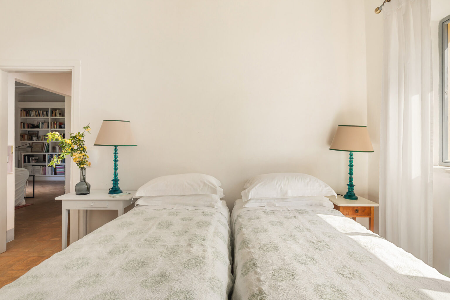 Cozy bedroom featuring two twin beds with floral bedding, turquoise bedside lamps, and a vase of yellow flowers on a side table, set against a bright, minimalist wall. | Only on SopranoVillas