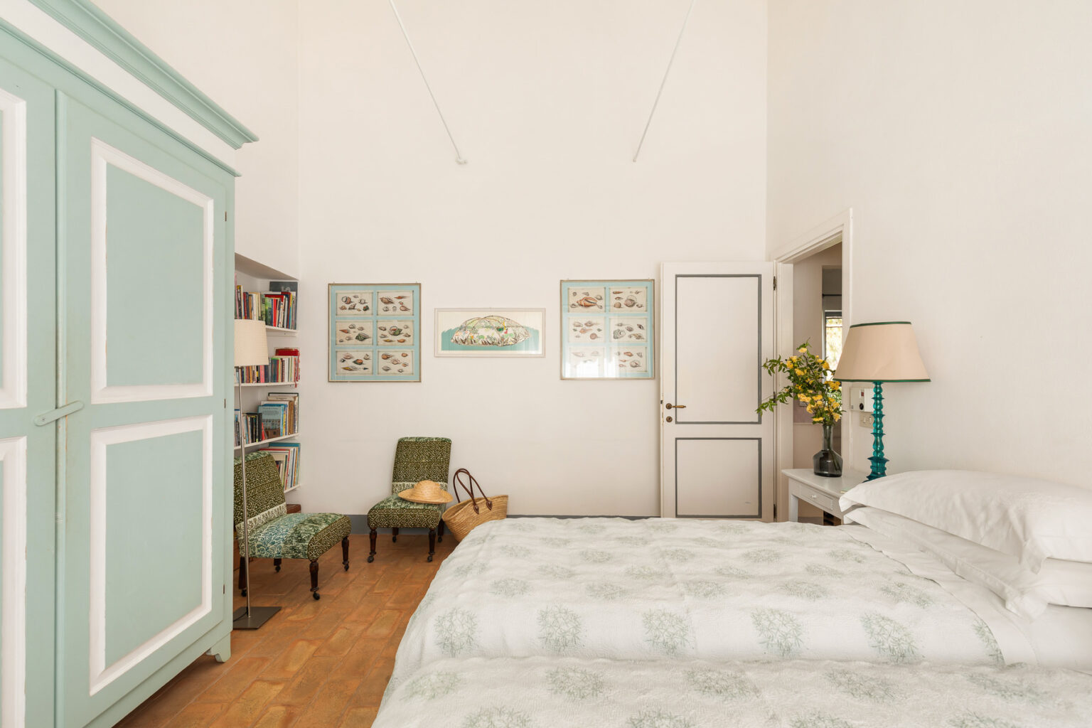 Cozy bedroom featuring a light color palette, a large bed with patterned bedding, a turquoise wardrobe, green accent chairs, and decorative wall art, creating a serene and inviting atmosphere. | Only on SopranoVillas
