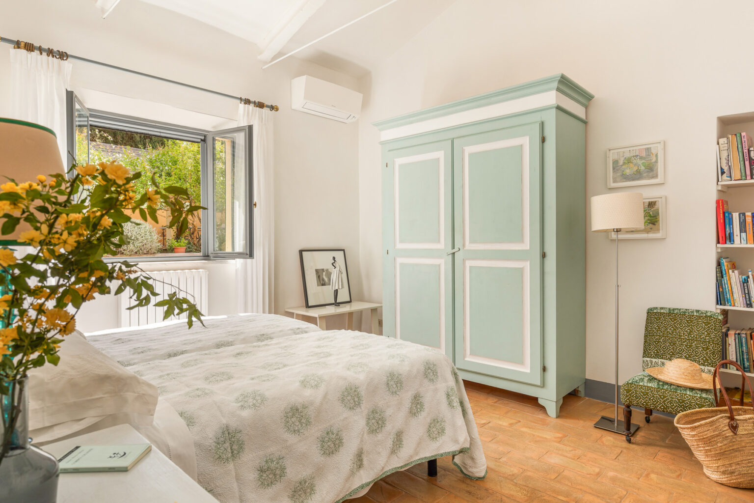 Bright and airy bedroom featuring a light green wardrobe, a cozy bed with a patterned blanket, and a reading nook with a chair and bookshelf. Natural light streams in through the window, enhancing the inviting atmosphere. | Only on SopranoVillas
