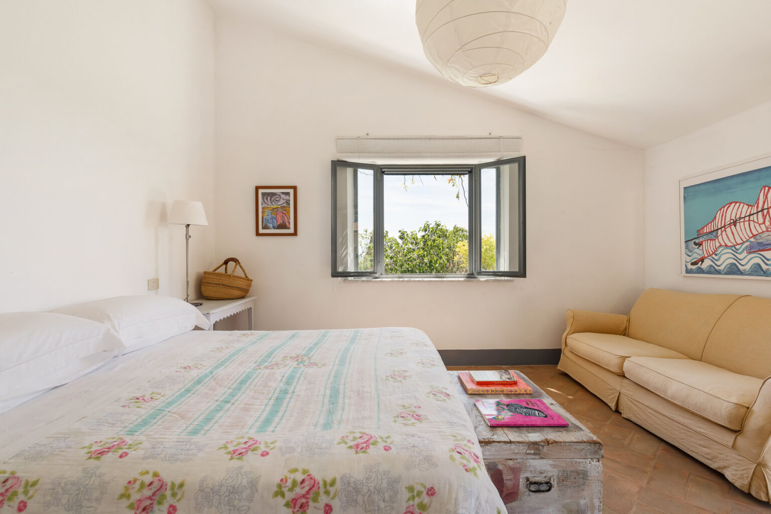 Bright and airy bedroom featuring a comfortable bed with a floral patterned quilt, a cozy sofa, and large windows offering a view of greenery. Decor includes a wall art piece and a side table with colorful magazines. | Only on SopranoVillas