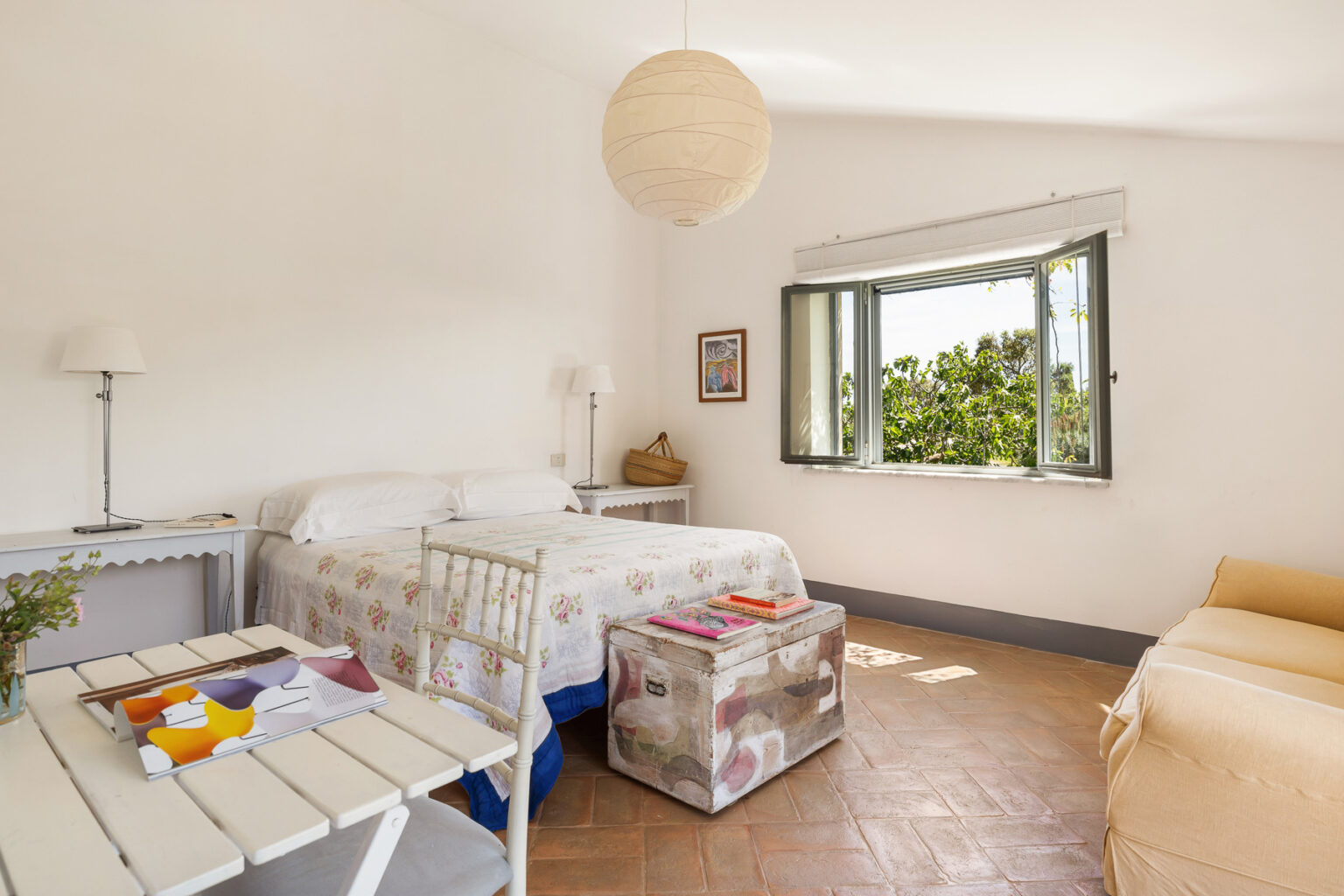 Bright and airy bedroom featuring a double bed, bedside table, and a cozy sitting area with natural light streaming in through a large window. Decorated with simple furnishings and a light color palette, creating a serene atmosphere. | Only on SopranoVillas