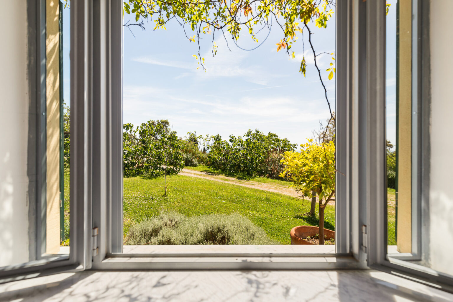 View from an open window showcasing a lush green garden with blooming trees and vibrant flowers under a clear blue sky. | Only on SopranoVillas