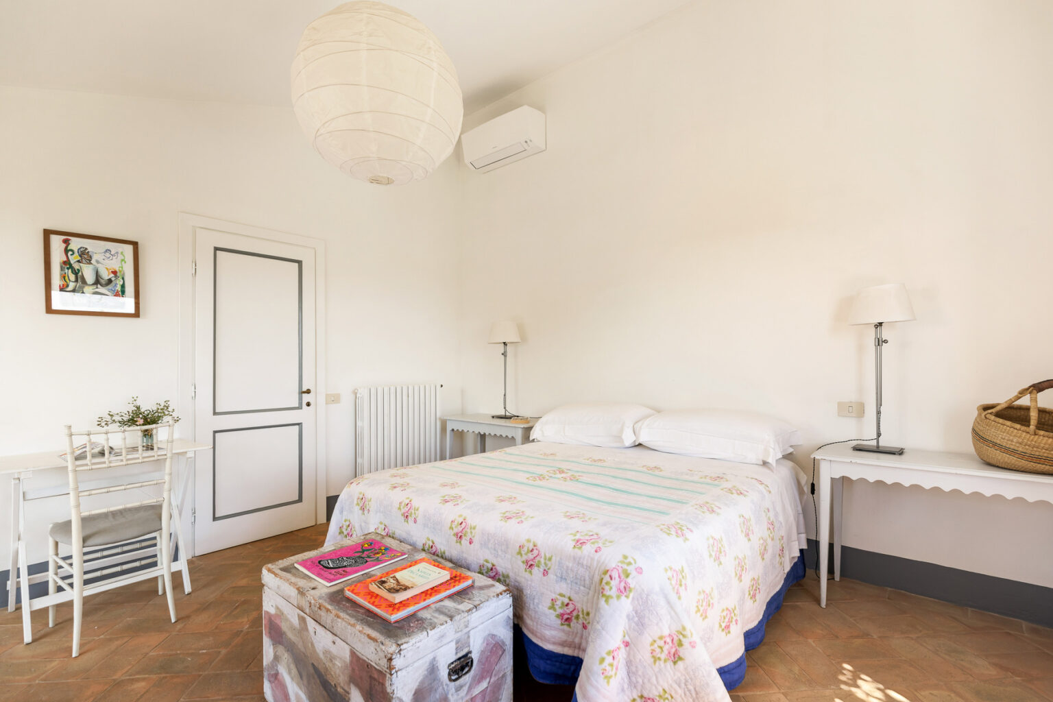 Cozy bedroom featuring a double bed with a floral-patterned quilt, a small wooden table, two chairs, a decorative lamp, and a light fixture. The room has white walls and wooden flooring, creating a bright and inviting atmosphere. | Only on SopranoVillas