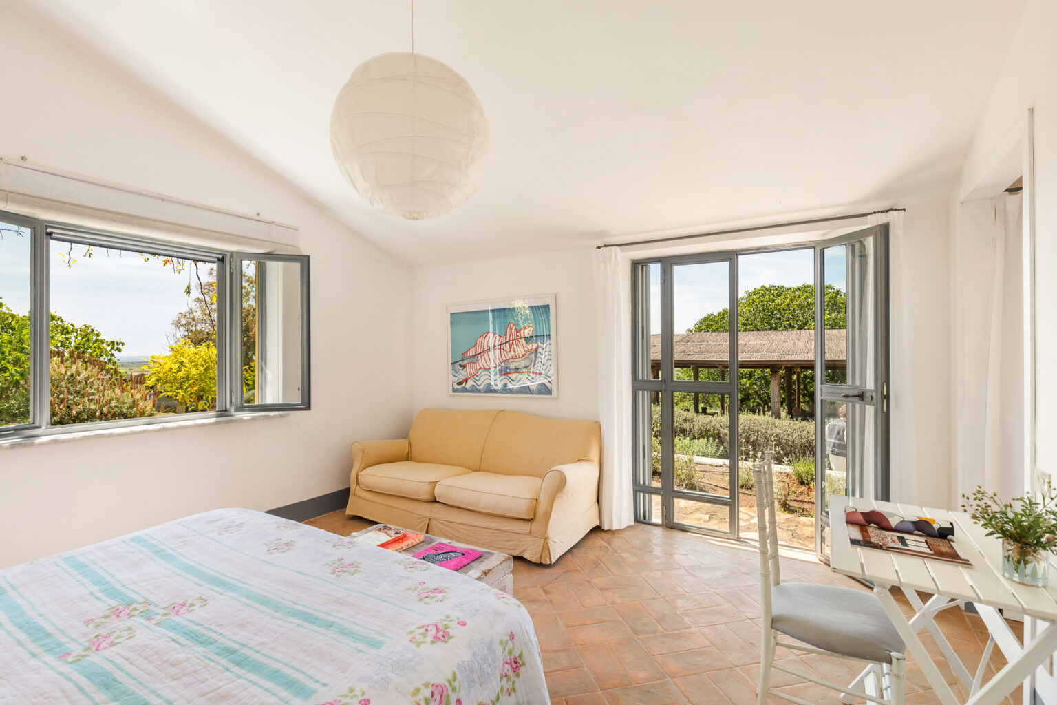 Bright and airy bedroom featuring a cozy sofa, large windows with garden views, and a dining table. Decorated with a colorful bedspread and artwork, this space offers a serene atmosphere for relaxation. | Only on SopranoVillas
