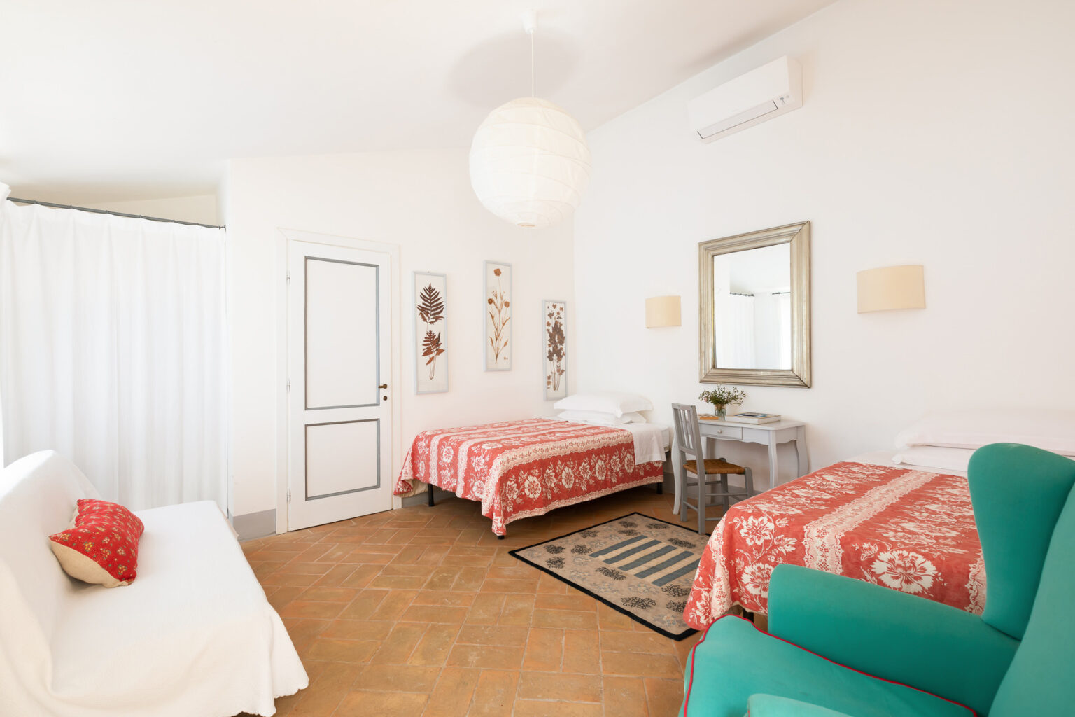Bright and airy bedroom featuring two beds with red patterned bedspreads, a green armchair, a small desk, and decorative wall art. The room includes large windows and a modern light fixture, creating a welcoming atmosphere. | Only on SopranoVillas
