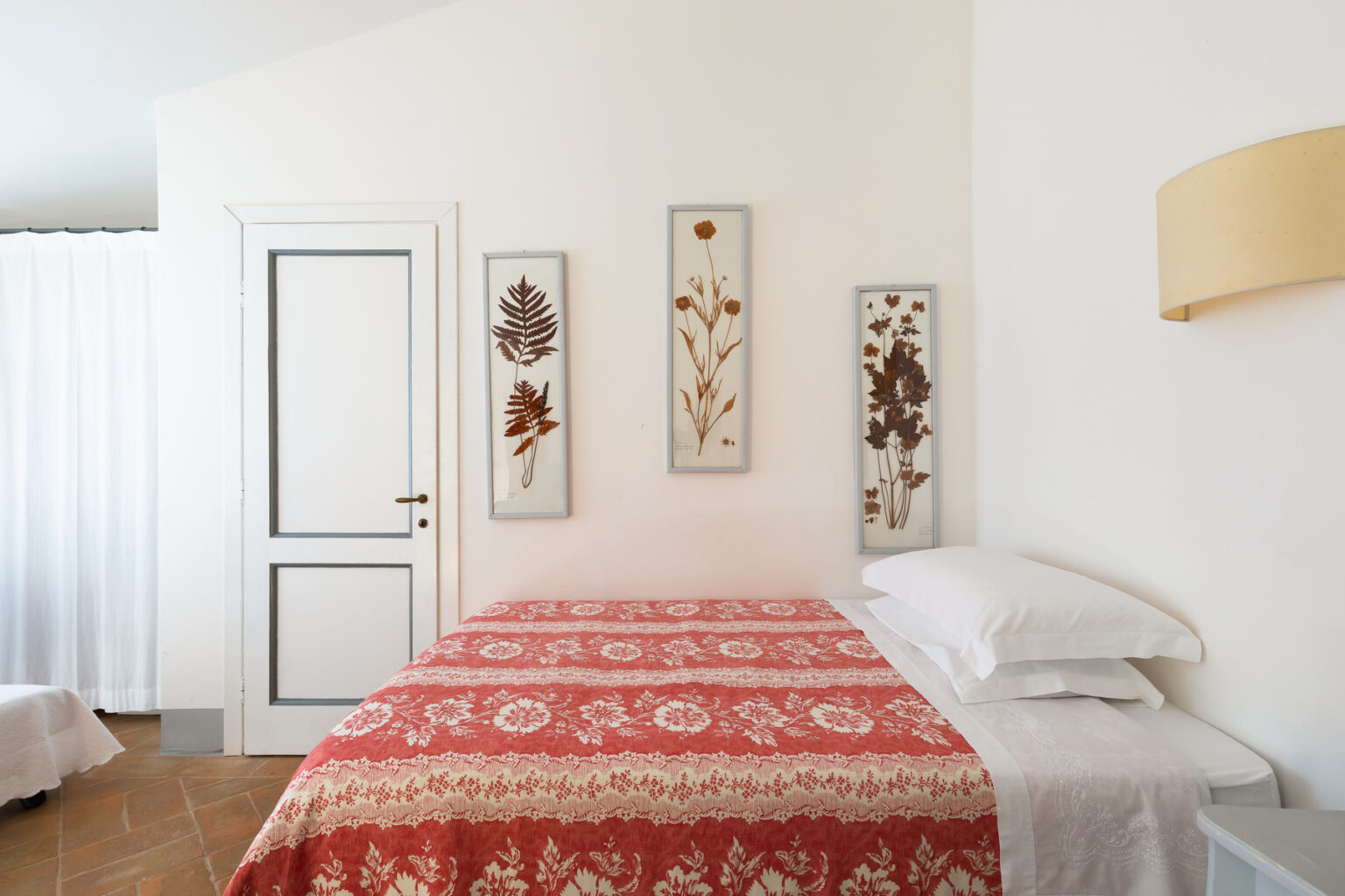 Cozy bedroom featuring a red patterned bedspread, white pillows, and framed dried flower artwork on the wall. A white door leads to another room, with soft natural light filtering through sheer curtains. Ideal for a serene and inviting atmosphere. | Only on SopranoVillas