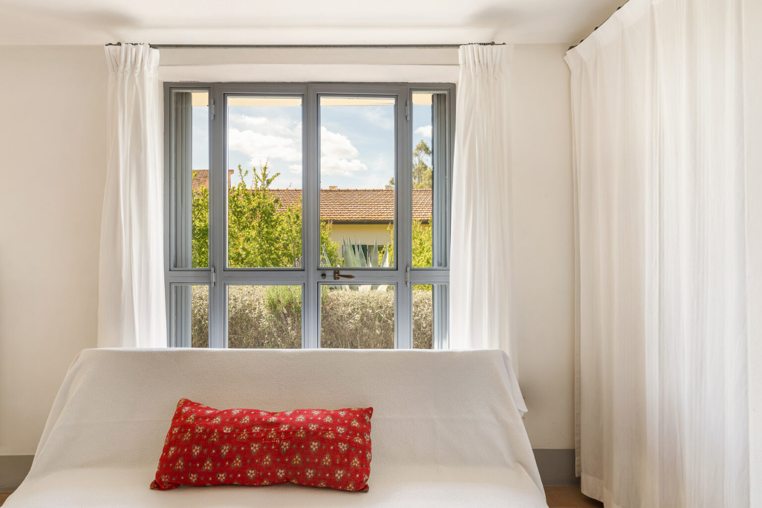 Cozy living room featuring a white sofa adorned with a red decorative pillow, large window with gray frames allowing natural light, and sheer white curtains, showcasing a view of greenery outside. | Only on SopranoVillas