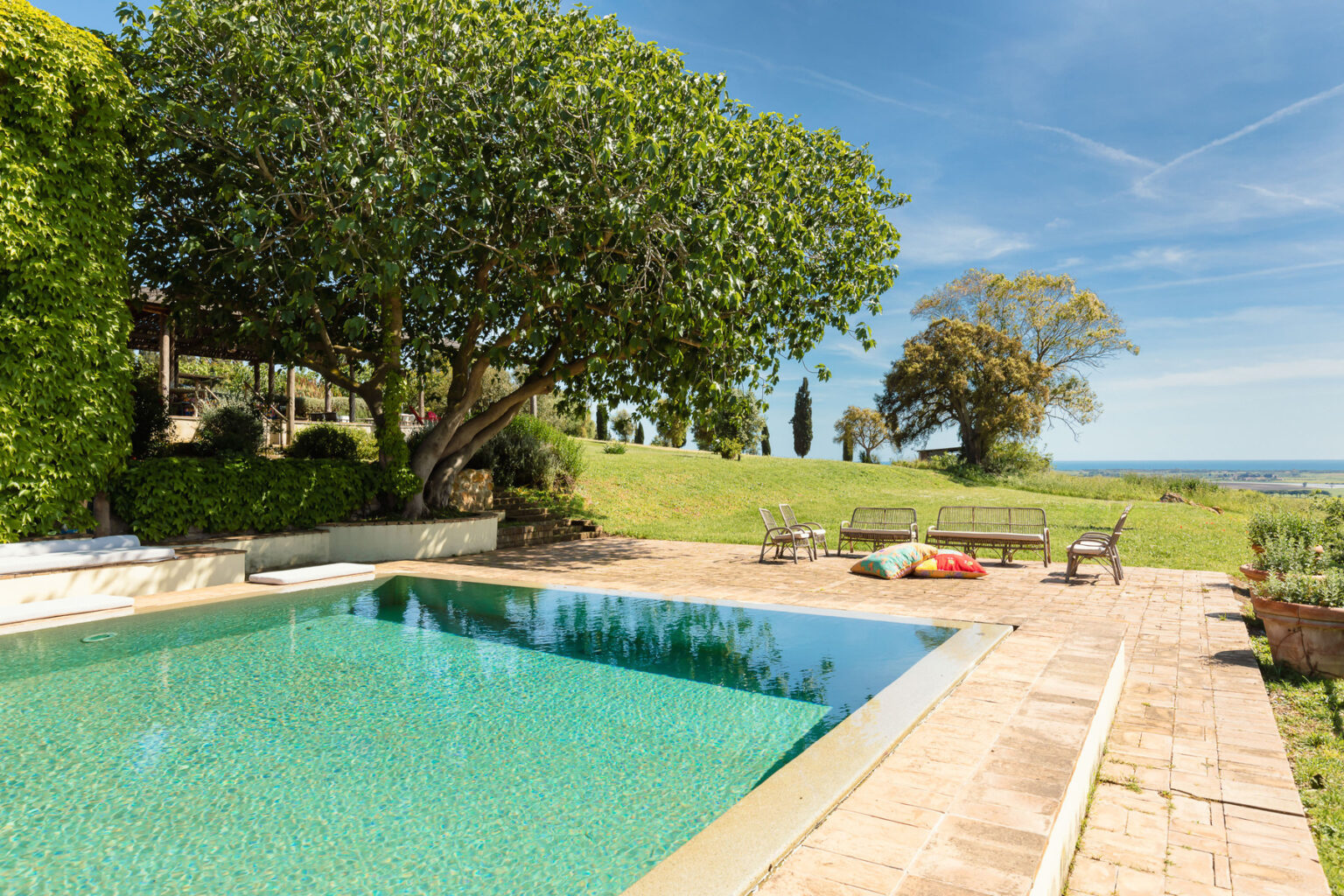 Serene outdoor pool area surrounded by lush greenery, featuring a large tree, comfortable seating, and a scenic view of the landscape under a clear blue sky. | Only on SopranoVillas