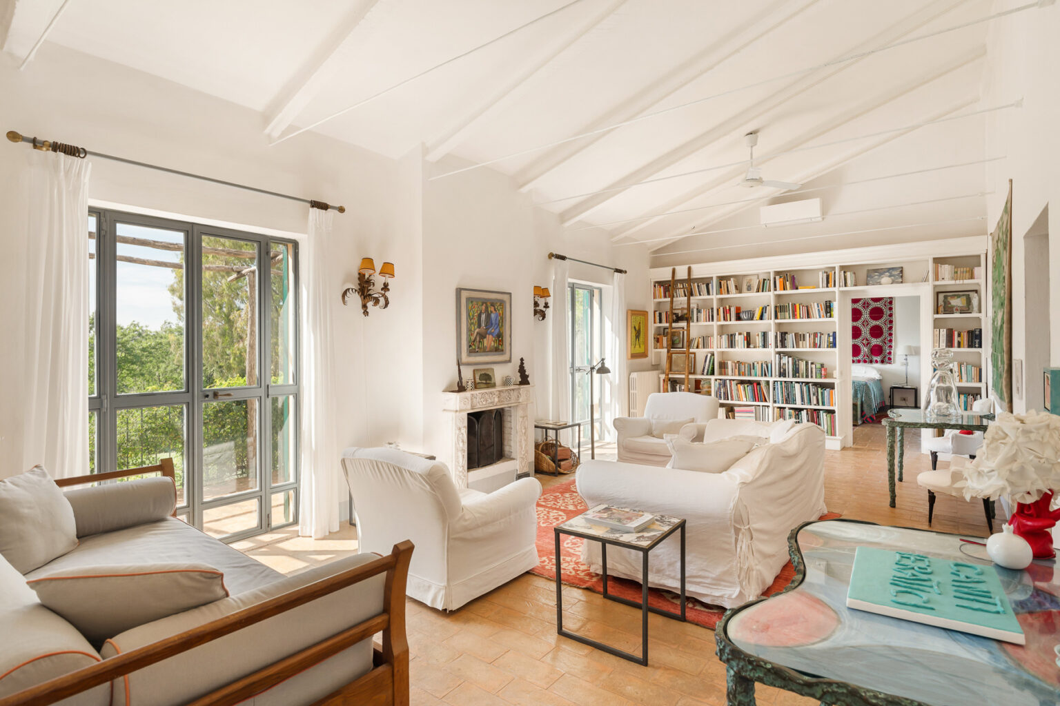 Bright and airy living room featuring white furniture, a cozy fireplace, and large windows with sheer curtains. A bookshelf filled with books adds character, while decorative artwork and a coffee table enhance the inviting atmosphere. The space is complemented by warm natural light and a view of greenery outside. | Only on SopranoVillas