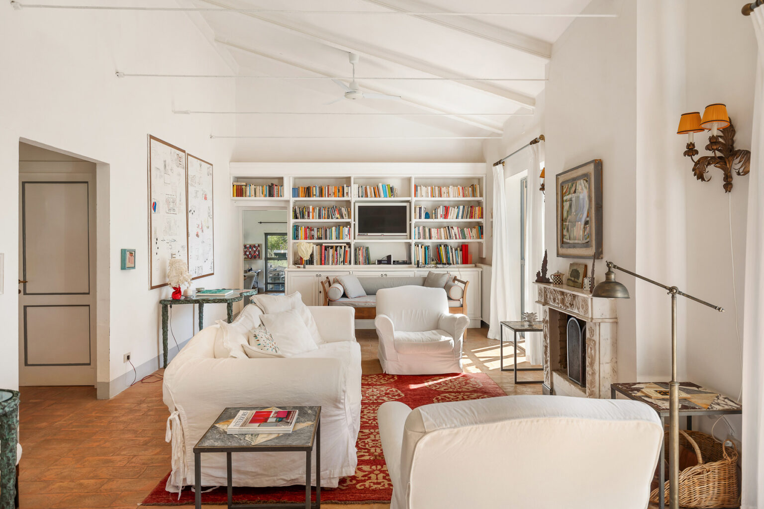 Bright and airy living room featuring white furniture, a cozy red area rug, and built-in bookshelves filled with books. The space includes a modern fireplace and large windows, creating an inviting atmosphere. | Only on SopranoVillas