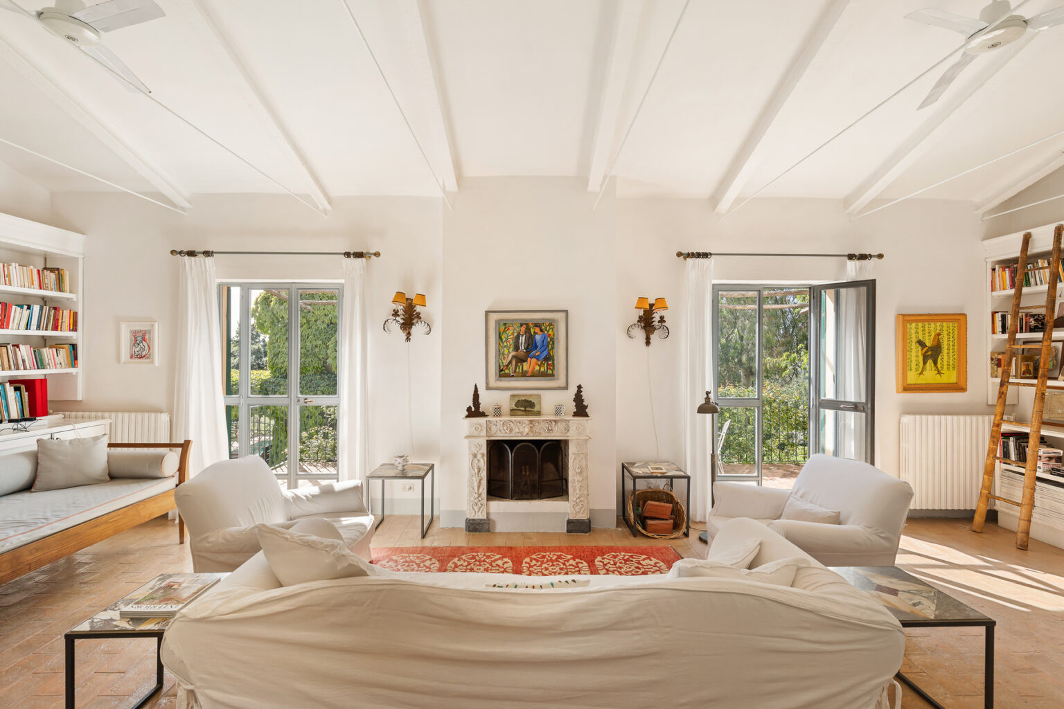 Cozy living room with white walls, wooden beams, and large windows allowing natural light. Features include a comfortable sofa, a decorative rug, a fireplace, and a bookshelf filled with books, creating an inviting atmosphere. | Only on SopranoVillas