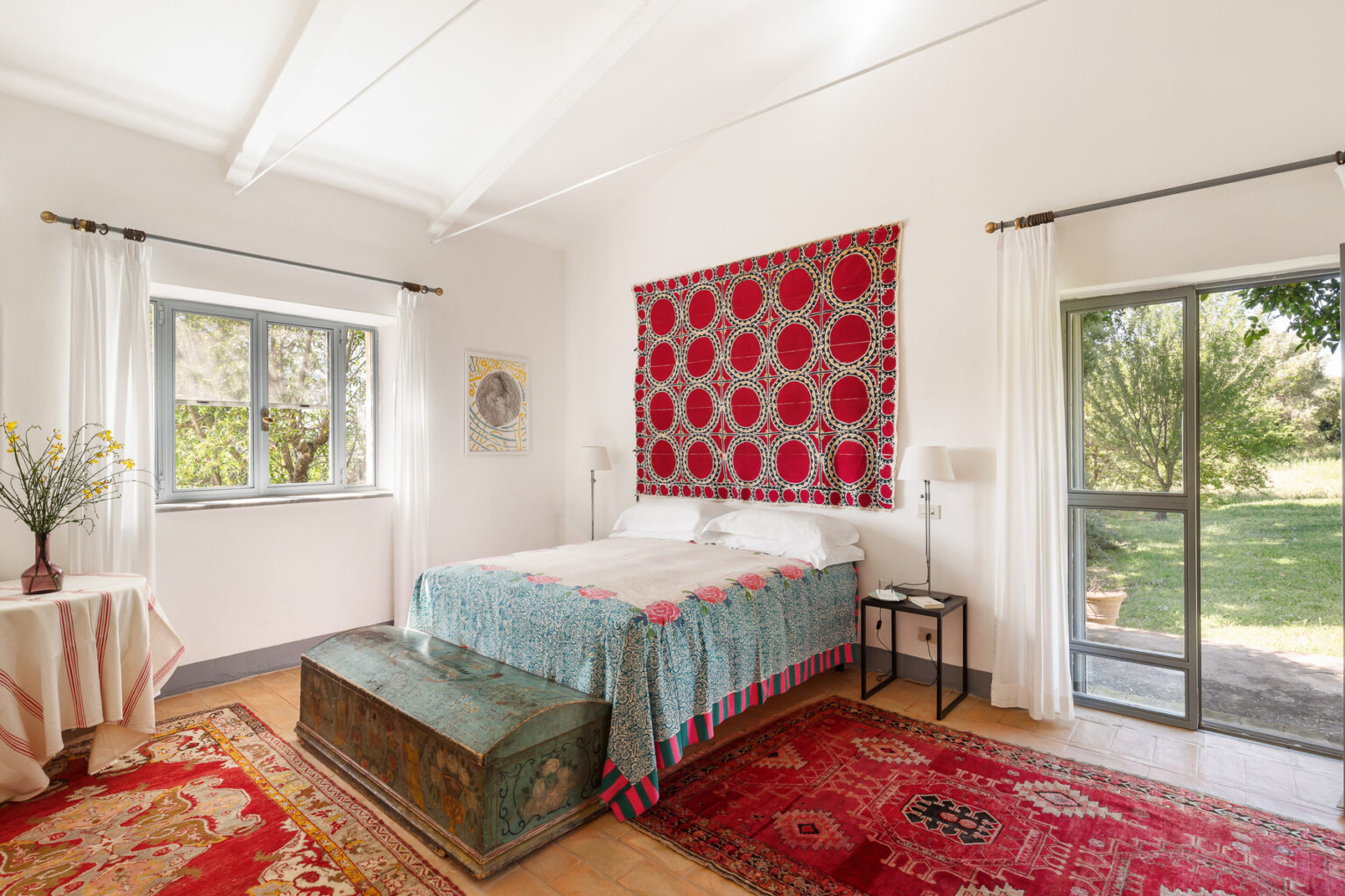 Cozy bedroom featuring a large bed with a colorful quilt, a decorative red wall hanging, and a wooden trunk at the foot of the bed. Natural light floods the room through large windows, complemented by elegant curtains and a small table with a vase of flowers. The space is adorned with vibrant rugs and opens to a green outdoor view. | Only on SopranoVillas