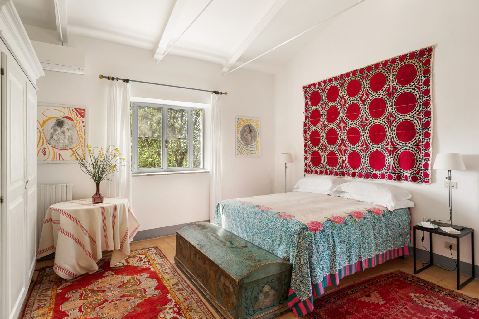 Cozy bedroom featuring a colorful patterned bedspread, vintage trunk at the foot of the bed, and decorative wall art. Natural light streams in through a window adorned with sheer curtains, enhancing the inviting atmosphere. | Only on SopranoVillas