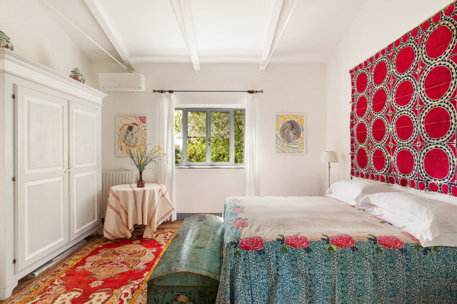 Bright and airy bedroom featuring a large bed with decorative bedding, a colorful patterned wall hanging, a vintage-style rug, and a small table with a vase of flowers. Natural light flows in through a window, enhancing the cozy atmosphere. | Only on SopranoVillas