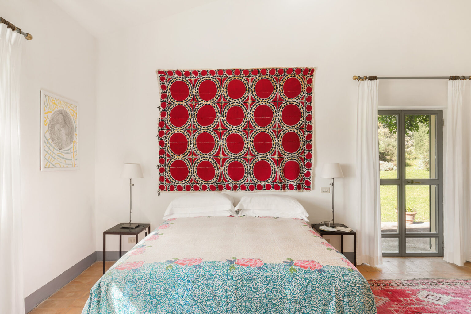 Cozy bedroom featuring a vibrant red wall tapestry, a neatly made bed with decorative bedding, bedside tables with lamps, and large windows allowing natural light to illuminate the space. | Only on SopranoVillas
