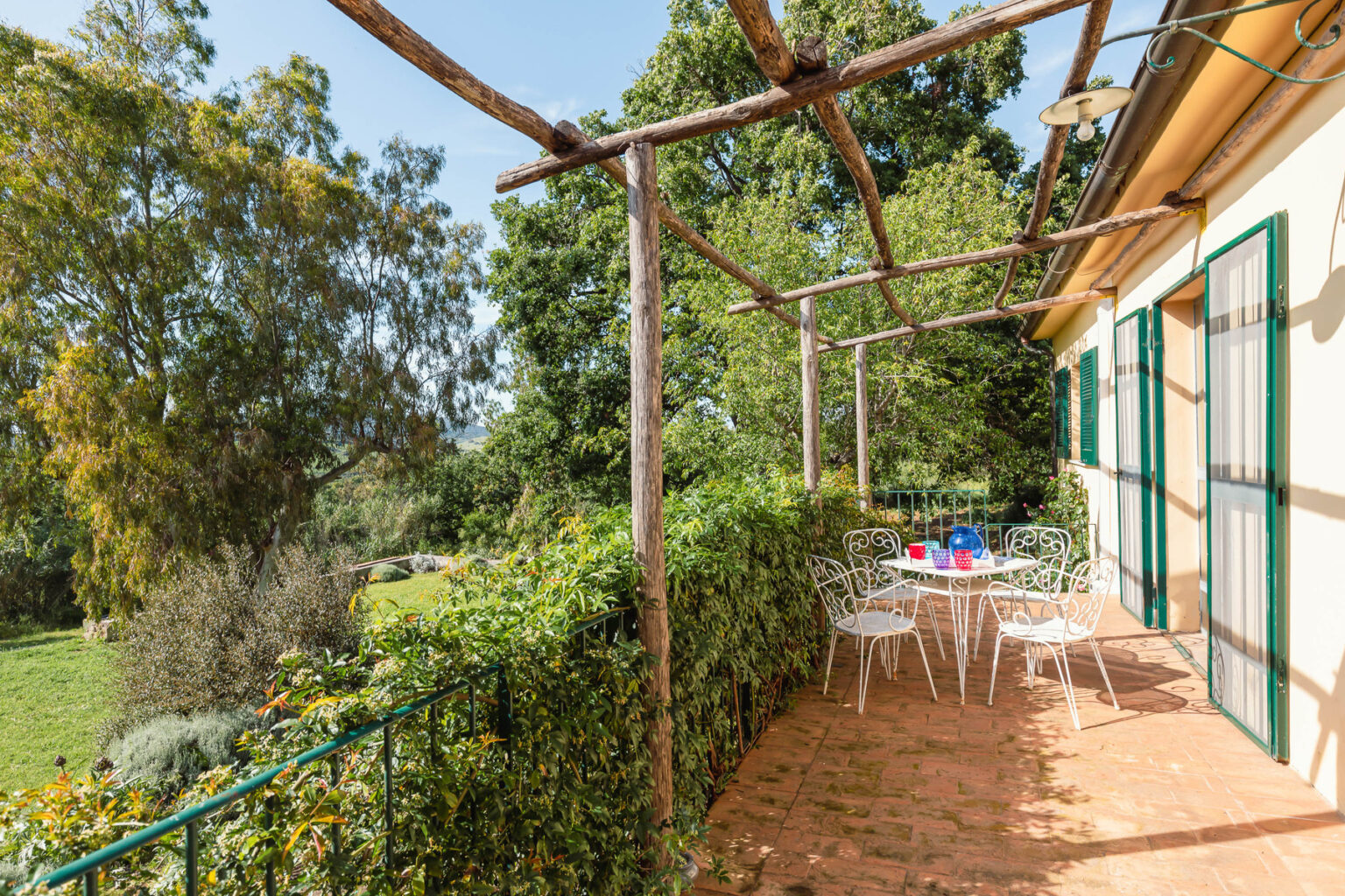 Outdoor patio area with a vine-covered pergola, featuring white chairs and a table set for dining, surrounded by lush greenery and trees. Ideal for relaxing in a serene garden setting. | Only on SopranoVillas