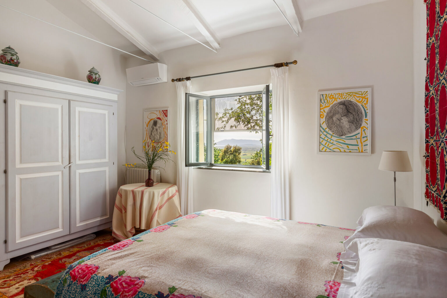 Bright and airy bedroom featuring white walls, a large window with a scenic view, a cozy bed, and decorative artwork. The room includes a stylish wardrobe and a table with a plant, showcasing a blend of comfort and modern design. | Only on SopranoVillas
