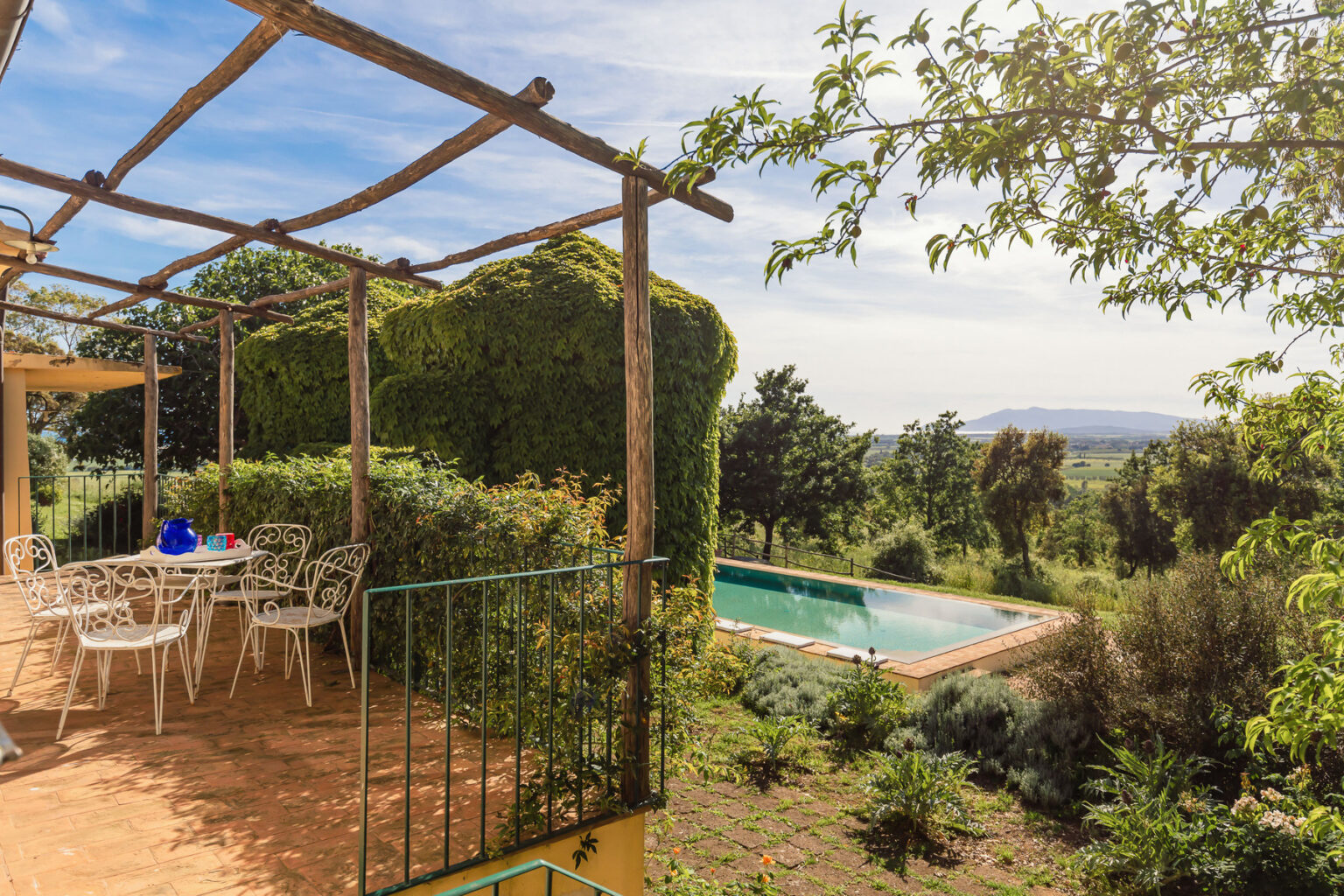 Scenic view of a terrace with a dining area overlooking a swimming pool surrounded by lush greenery and distant hills. Ideal for outdoor relaxation and entertaining. | Only on SopranoVillas