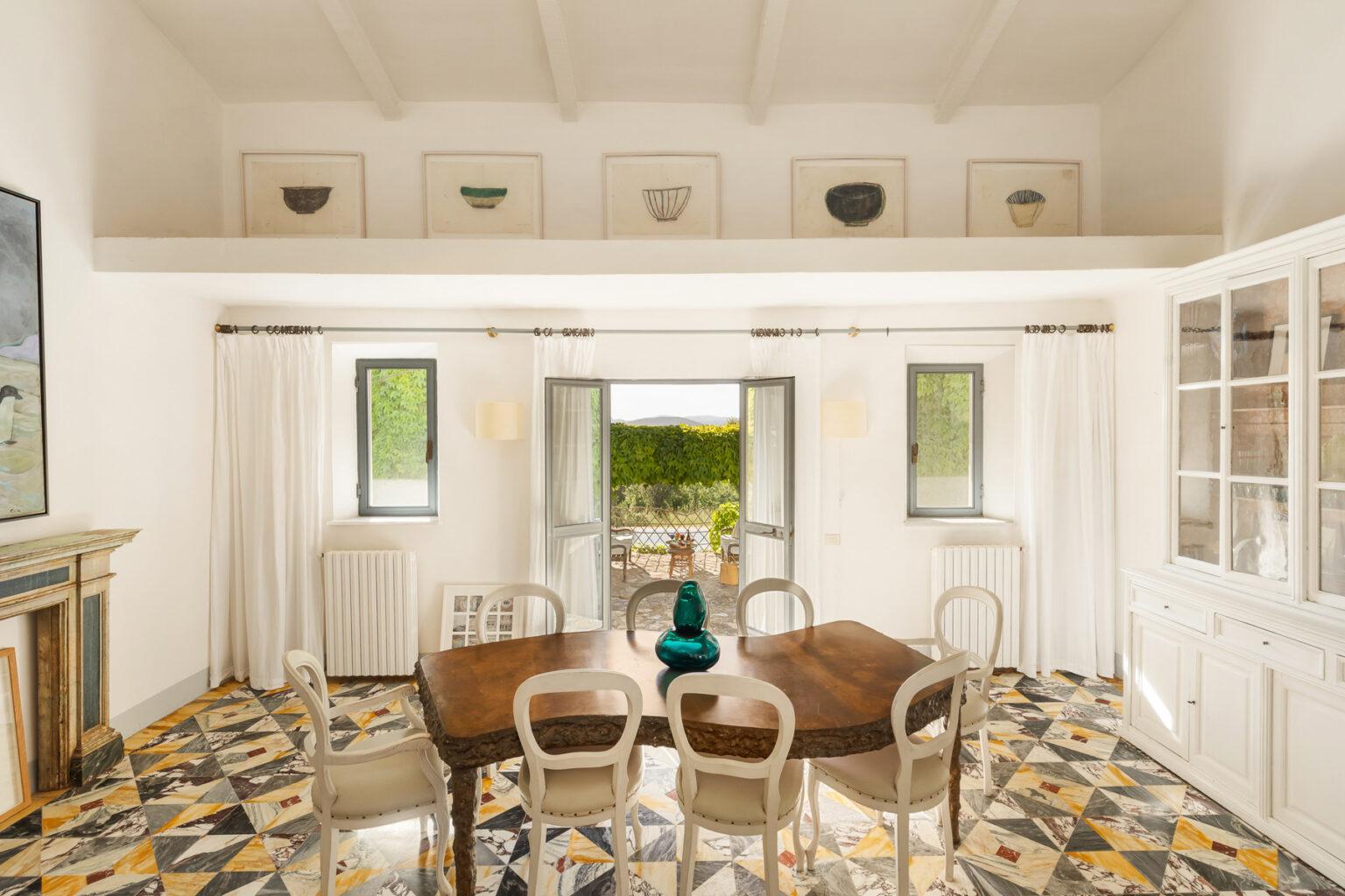 Bright and airy dining room featuring a wooden table surrounded by elegant chairs, large windows with garden views, and decorative art pieces on the walls. The room has a stylish tiled floor and a cozy ambiance. | Only on SopranoVillas