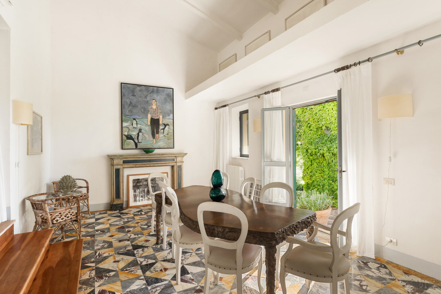 Bright and airy dining room featuring a wooden table surrounded by elegant chairs, a striking artwork on the wall, and a view of lush greenery through an open window. The space showcases unique patterned flooring and a stylish decorative element on the table. | Only on SopranoVillas