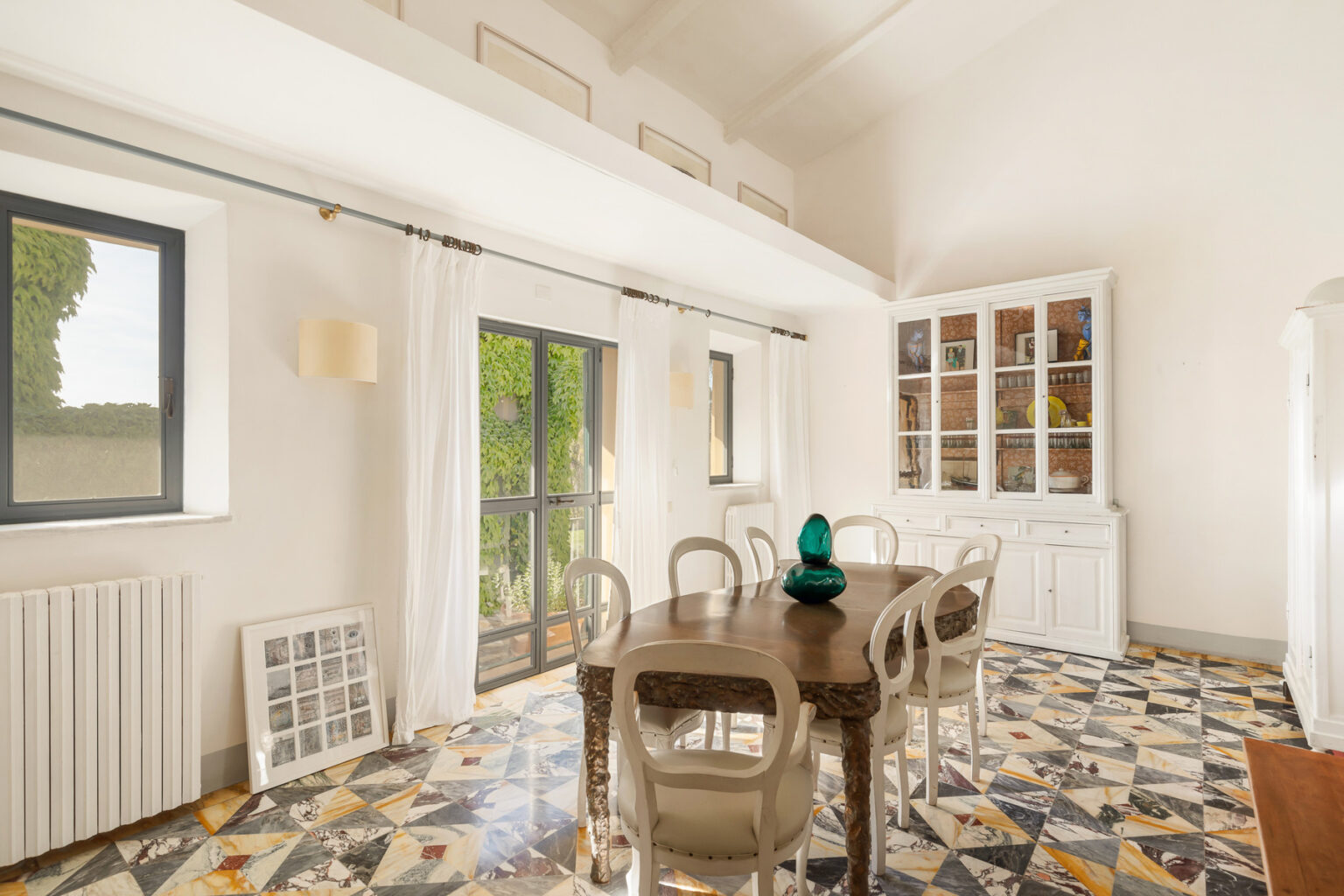 Bright and airy dining room featuring a wooden table surrounded by stylish chairs, a decorative green glass centerpiece, large windows with sheer curtains, and a white cabinet displaying colorful decor. The floor showcases a unique patterned tile design, enhancing the room's modern aesthetic. | Only on SopranoVillas
